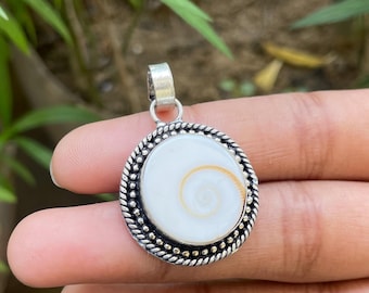 May include: A round pendant with a white center and a spiral design in shades of gold. The pendant is set in a silver-colored metal frame with a textured border. The bail is also silver-colored. The pendant is held in a hand.