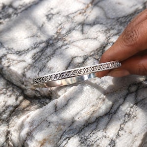 Può includere: Un braccialetto rigido d'argento con un intricato motivo ripetuto di piccole forme floreali o a foglia stilizzate. Il braccialetto è presentato su una superficie di pietra strutturata, grigia e bianca, che ne mette in risalto il design e la lucentezza metallica.