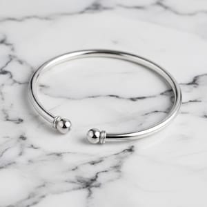 May include: A silver-colored open cuff bracelet with a smooth, polished finish. The bracelet has rounded ball-shaped ends. The jewelry is displayed on a white and gray marble surface.