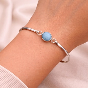 May include: A silver-toned bangle bracelet featuring a round, light blue gemstone. The bracelet has a simple, elegant design with a smooth, polished finish. The gemstone is set in a bezel setting and is the focal point of the bracelet.