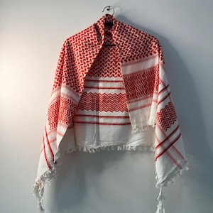 Pode incluir: Um lenço keffiyeh vermelho e branco com um padrão geométrico xadrez tradicional, faixas horizontais listradas e borlas brancas decorativas ao longo da bainha inferior. O tecido está drapeado e pendurado num cabide de metal.