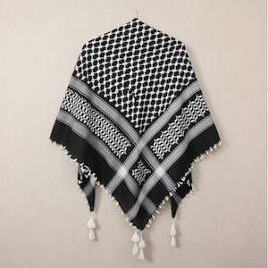 Può includere: Una sciarpa keffiyeh bianca e nera con un motivo geometrico e nappe decorative. La sciarpa è di forma quadrata e appesa a un gancio di metallo. Il design include un motivo ripetuto di forme bianche e nere, con un bordo di strisce grigie e bianche.