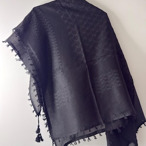 Puede incluir: Un pañuelo negro con un patrón geométrico tejido en tono sobre tono en toda la tela. El accesorio de forma cuadrada está rematado con borlas decorativas anudadas a lo largo de los bordes y se muestra colgado de un gancho de metal.