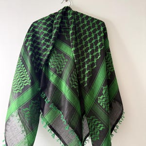 May include: A square scarf featuring a black and bright green checkered pattern with a repeating leaf-like motif. The fabric has a woven texture and is finished with small green tassels along the edges, hanging from a metal hanger.