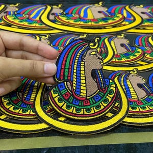 Może przedstawiać: Haftowana naszywka przedstawiająca profil egipskiego faraona z kolorowym nakryciem głowy. Naszywka ma żółtą obwódkę i jest umieszczona na czarnym tle. Obraz przedstawia kilka naszywek.