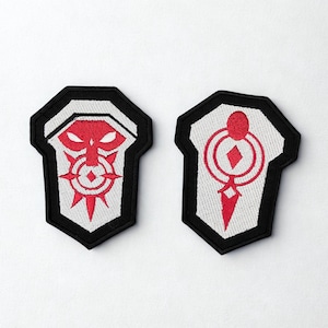 May include: Two embroidered patches with a shield shape. Each patch has a white background with a red design. One features a face-like design, the other a geometric pattern. Both have a black border.