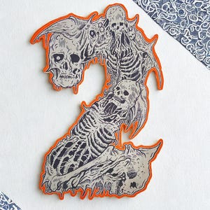 Peut inclure: Un autocollant découpé en forme de chiffre 2, avec une illustration détaillée en noir et blanc de crânes et de squelettes, bordée d'une bordure orange. Le design a un thème gothique ou d'horreur.