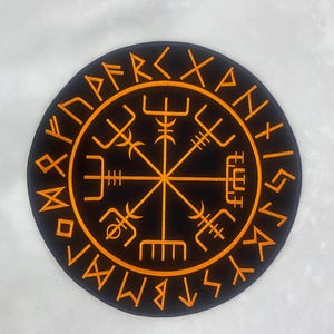 Könnte beinhalten: Ein schwarzes, kreisförmiges Design mit einem gold-orangenen Vegvisir-Symbol, einem Wikingerkompass, umgeben von Runeninschriften. Das Design ist auf einem hellgrauen Hintergrund zentriert und erzeugt einen auffälligen Kontrast.