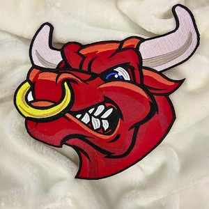 Könnte beinhalten: Ein roter Bullenkopf-Aufnäher mit gelbem Nasenring und weißen Hörnern. Der Bulle hat einen grimmigen Ausdruck mit weißen Zähnen und blauen Augen. Der Aufnäher ist mit schwarzen Konturen bestickt.
