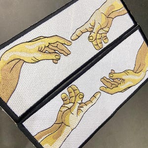 May include: Two rectangular patches featuring an embroidered depiction of Michelangelo's 'The Creation of Adam' from the Sistine Chapel ceiling. The patches show two hands reaching towards each other, with yellow and tan thread on a white background.
