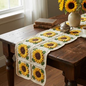 May include: A handmade crochet table runner featuring a repeating sunflower design. The sunflowers have yellow petals and brown centers, with green leaves. The runner is displayed on a dark wooden table, with a vase of sunflowers, books, and a teacup.
