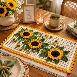 May include: A crocheted table runner with a sunflower design. The runner has a white grid pattern with a yellow scalloped border. It features three large sunflowers with brown centres, yellow petals, and green leaves. A vase of sunflowers and a framed sign are also visible.