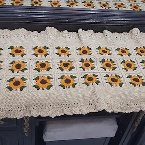 Könnte beinhalten: Cremefarbener, ovaler Häkeltischläufer mit einem sich wiederholenden Muster aus Sonnenblumen. Jede Sonnenblume hat gelbe Blütenblätter und eine braune Mitte. Der Läufer hat einen gewellten Rand.