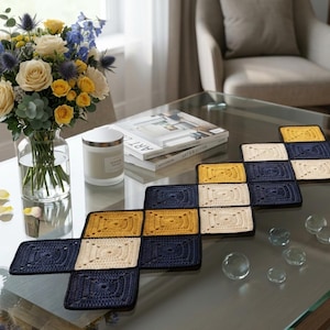 May include: A crocheted table runner in a geometric pattern of navy blue, cream, and yellow squares. The runner is displayed on a glass coffee table, alongside a vase of flowers and a candle.