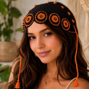 May include: A black crochet headpiece featuring a row of circular orange and black motifs across the front. The accessory includes small orange tassels along the top edge and two long, braided orange ties hanging down the sides with tassels.