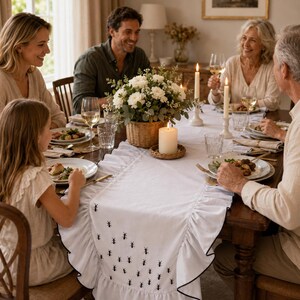 May include: A white rectangular table runner featuring a ruffled edge with a thin black trim and a repeating pattern of small black embroidered ants. The runner is laid across a wooden dining table set with plates, glasses, and candles.
