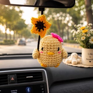 May include: A handmade crochet car charm featuring a yellow chick with a pink bow and a sunflower. The chick is holding the sunflower with a black stem. The charm is hanging from a car's rearview mirror.