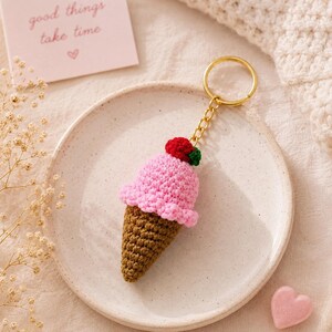 May include: A crochet ice cream cone keychain featuring a brown waffle cone, a pink scoop of ice cream, and a small red cherry with a green stem on top. The keychain is attached to a gold-toned metal ring and chain, resting on a speckled plate.