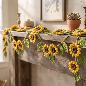 May include: A decorative garland featuring crocheted sunflowers with bright yellow petals and dark brown centers, interspersed with green crocheted leaves. The floral garland is draped across a surface, showcasing multiple connected flower heads.