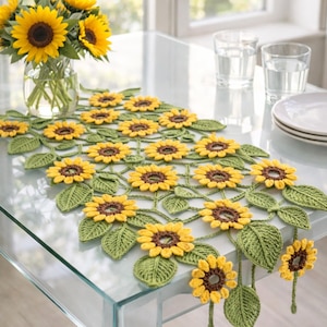 May include: A crocheted sunflower table runner featuring yellow flower heads and green leaves. The runner is displayed on a glass table. A vase of sunflowers and plates are in the background. The runner is a decorative item.