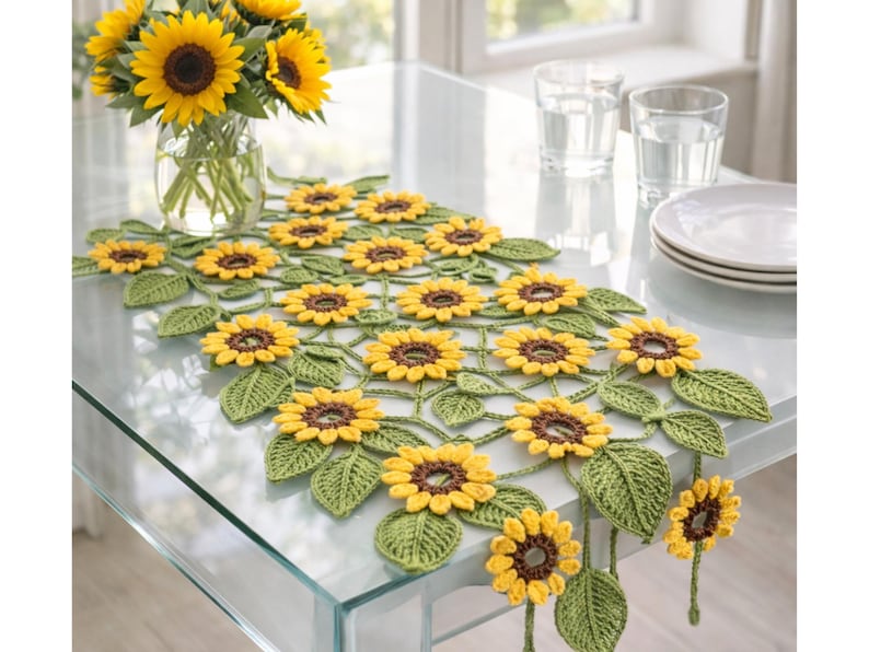 May include: A crochet table runner featuring a repeating pattern of yellow sunflowers with dark brown centers and textured green leaves. The handcrafted textile piece is arranged in a lattice design with several sunflower motifs extending over the edge.