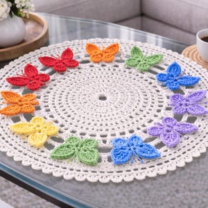 Könnte beinhalten: Eine cremefarbene, gehäkelte Tischdecke mit gewelltem Rand, verziert mit zwölf bunten gehäkelten Schmetterlingen. Die Schmetterlinge sind in den Farbtönen Rot, Orange, Gelb, Grün, Blau und Lila gehalten. Eine Tasse Kaffee steht in der Nähe.