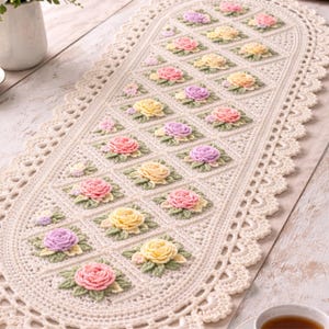 May include: Cream-colored, oval-shaped crochet table runner with a floral design. The runner features a repeating pattern of pink, yellow, and purple roses within diamond-shaped blocks, surrounded by a scalloped edge. The runner is displayed on a light-colored wooden table.