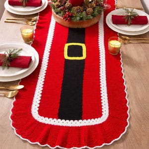 May include: A festive red and white crocheted table runner with a black belt design and a yellow buckle. The runner is decorated with a white scalloped edge. A centerpiece with a red candle and greenery sits on the runner, with place settings on either side.
