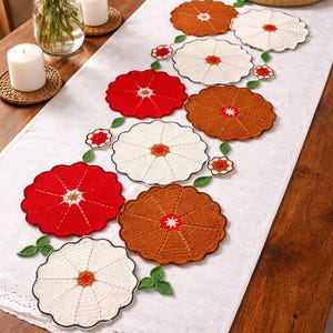 May include: A white table runner with a floral crochet design in red, cream, and brown. The runner is decorated with crocheted flowers and green leaves, creating a decorative centerpiece for a dining table.