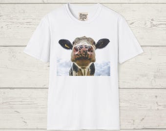 May include: White t-shirt featuring a close-up image of a cow's face. The cow has a pink and black nose, black ears, and a tag on one ear. The background is a cloudy sky. The shirt has a small tag near the neckline.
