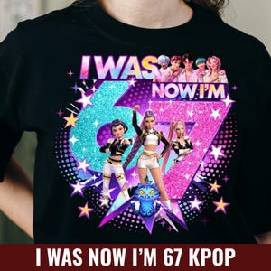 May include: Black t-shirt with a graphic design featuring the text "I WAS NOW I'M 67 KPOP". The design includes illustrations of K-pop group members and the number 67 in a glittery style, with stars and a blue cartoon cat.