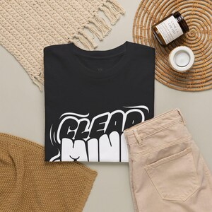 May include: A black crew neck t-shirt featuring the white text "CLEAR MIND" in a bold, stylized bubble font. The shirt is folded and displayed alongside a pair of light beige denim pants, a textured cream-colored fabric, and a small candle.