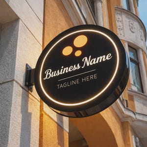 Może przedstawiać: Okrągły, czarny szyld firmowy z jasną, białą obwódką i napisem "Business Name" w eleganckiej czcionce. Poniżej wyświetlany jest tekst "TAGLINE HERE". Znak posiada trzy pomarańczowe okręgi i jest zamontowany na budynku.