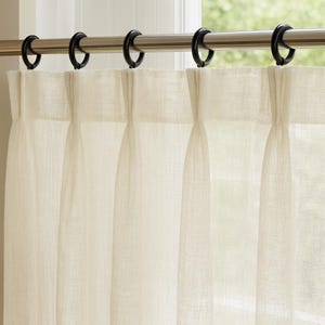 May include: Cream-colored linen curtains hanging from a silver rod with black metal rings. The curtains are pleated and hang in vertical folds, with natural light filtering through the sheer fabric. The window is partially visible in the background.