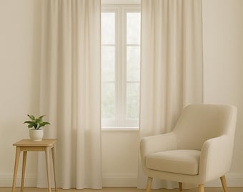 May include: A light-toned living room with cream curtains and a window. A small wooden side table with a plant is on the left, and a cream armchair is on the right. The room has a minimalist design, with a neutral colour palette.