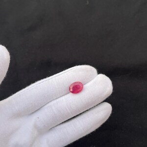 Puede incluir: Una gema de rubí facetada de forma ovalada. El rubí es de color rojo intenso y se sostiene en una mano enguantada de blanco sobre un fondo negro. La gema es una piedra preciosa.