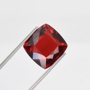 Pode incluir: Uma pedra preciosa vermelha escura, lapidada em forma quadrada, é segurada por uma pinça prateada sobre um fundo branco. As múltiplas facetas da gema refletem a luz, criando um brilho brilhante. A cor da gema é um vermelho rico e saturado.