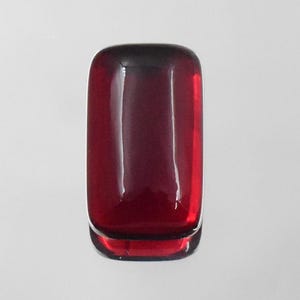 May include: A rectangular cabochon gemstone with a deep, translucent red colour and a smooth, polished surface. The stone features rounded corners and a high-gloss finish that reflects light, showcasing its rich, saturated crimson hue.