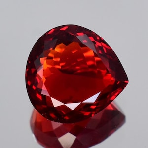 May include: A pear-shaped faceted gemstone with a deep red color and orange undertones. The stone features a brilliant cut with multiple triangular and kite-shaped facets that reflect light, creating a vibrant display of color and internal clarity.
