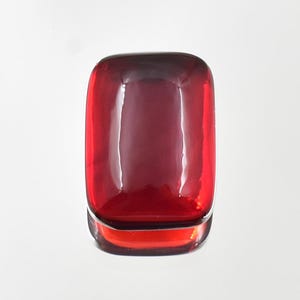 May include: A rectangular, deep red gemstone with a smooth, polished cabochon surface. The stone features rounded corners and a translucent quality that allows light to pass through, creating a bright reflection on its top curved face.
