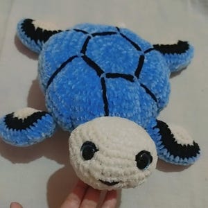 May include: A handmade plush turtle toy. The turtle has a blue shell with black accents, a white head, and black eyes. The flippers are blue with black and white details. The toy is made of soft, textured yarn.