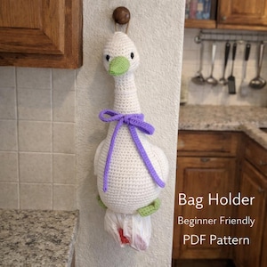 Puede incluir: Un soporte para bolsas de ganchillo con forma de ganso blanco con un lazo morado y detalles verdes. El ganso tiene un pico verde y ojos negros. El soporte para bolsas está colgado en una pared. El texto en la imagen dice "Bag Holder Beginner Friendly PDF Pattern."