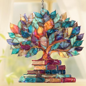 May include: A colorful, stained-glass style tree ornament. The tree has a brown trunk and branches with vibrant leaves in shades of blue, purple, orange, and yellow. The tree sits atop a stack of books. A chain is attached for hanging.