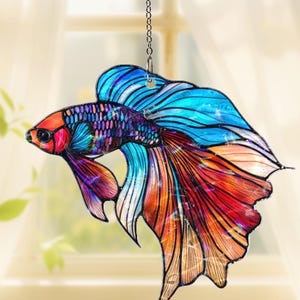 May include: A colorful, stained-glass-style betta fish ornament. The fish features vibrant hues of red, orange, blue, and purple. It is suspended by a chain, suitable for hanging as a decorative piece. The fish's fins and tail are detailed.