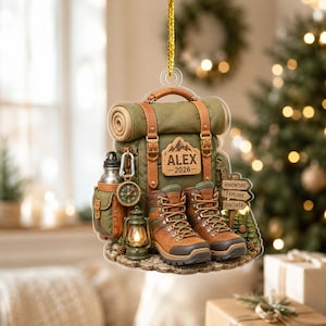 May include: A hanging ornament featuring an olive green backpack with brown straps, a pair of brown hiking boots, a compass, a water bottle, and a lantern. A wooden sign reads ALEX 2026, and a directional sign reads ADVENTURE, EXPLORE, DISCOVER.