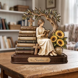 May include: A decorative figurine featuring a person reading a book while seated beside a stack of eight books labeled with religious text, an olive tree, a small vase with three sunflowers, and two crosses, all set on a base with the name Sophia.