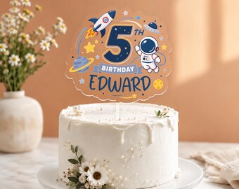 Puede incluir: Un pastel blanco glaseado con un adorno para el 5º cumpleaños. El adorno presenta un cohete, un astronauta y el nombre "Edward". El pastel está decorado con flores blancas y perlas.