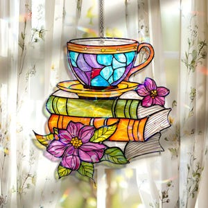 May include: A stained glass suncatcher featuring a teacup and saucer atop a stack of books, with flowers and leaves. The teacup is blue, purple, and orange. The books are green and orange. The flowers are pink and purple.