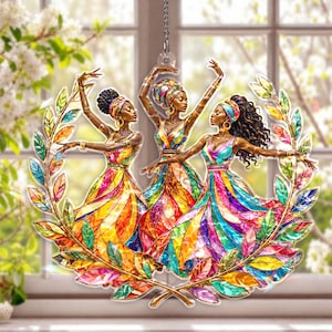 May include: A decorative ornament featuring three women in colorful dresses, framed by a gold-colored laurel wreath. The women are depicted with their arms raised, wearing head wraps and jewelry. The dresses are a vibrant mix of colors, including yellow, red, blue, and green.