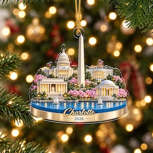 May include: A flat, circular hanging ornament featuring a detailed illustration of Washington D.C. landmarks, including the Capitol, Washington Monument, and White House, surrounded by pink cherry blossoms, blue water, and the text Charlotte 2026.